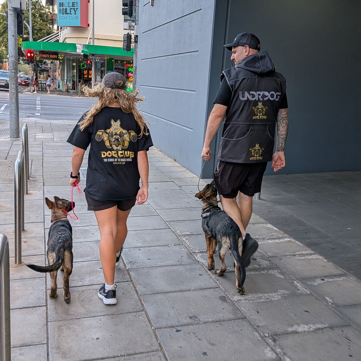 Two people walking dogs on a city sidewalk with a building and street signs in the background.