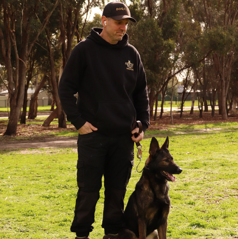 Man in black hoodie and pants with a dog on a leash in a park.